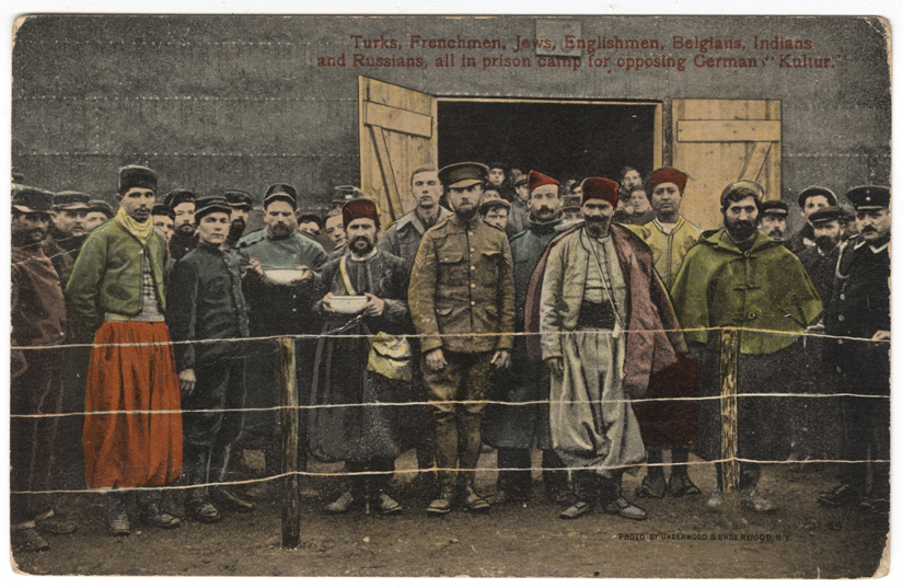 Colorized photograph of a large group of men standing behind barbed wire looking at the viewer. They are dressed in a variety of national clothing.