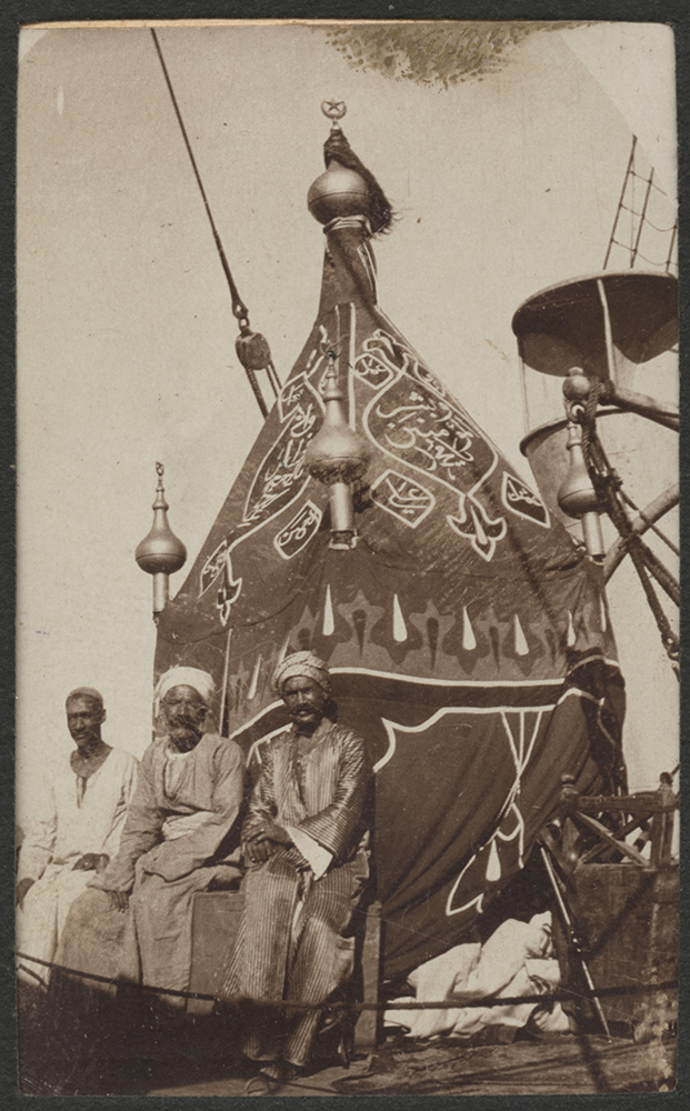 Sepia photograph of three brown-skinned men wearing loose robes sitting in front of a tent-like structure painted or embroidered with intricate designs