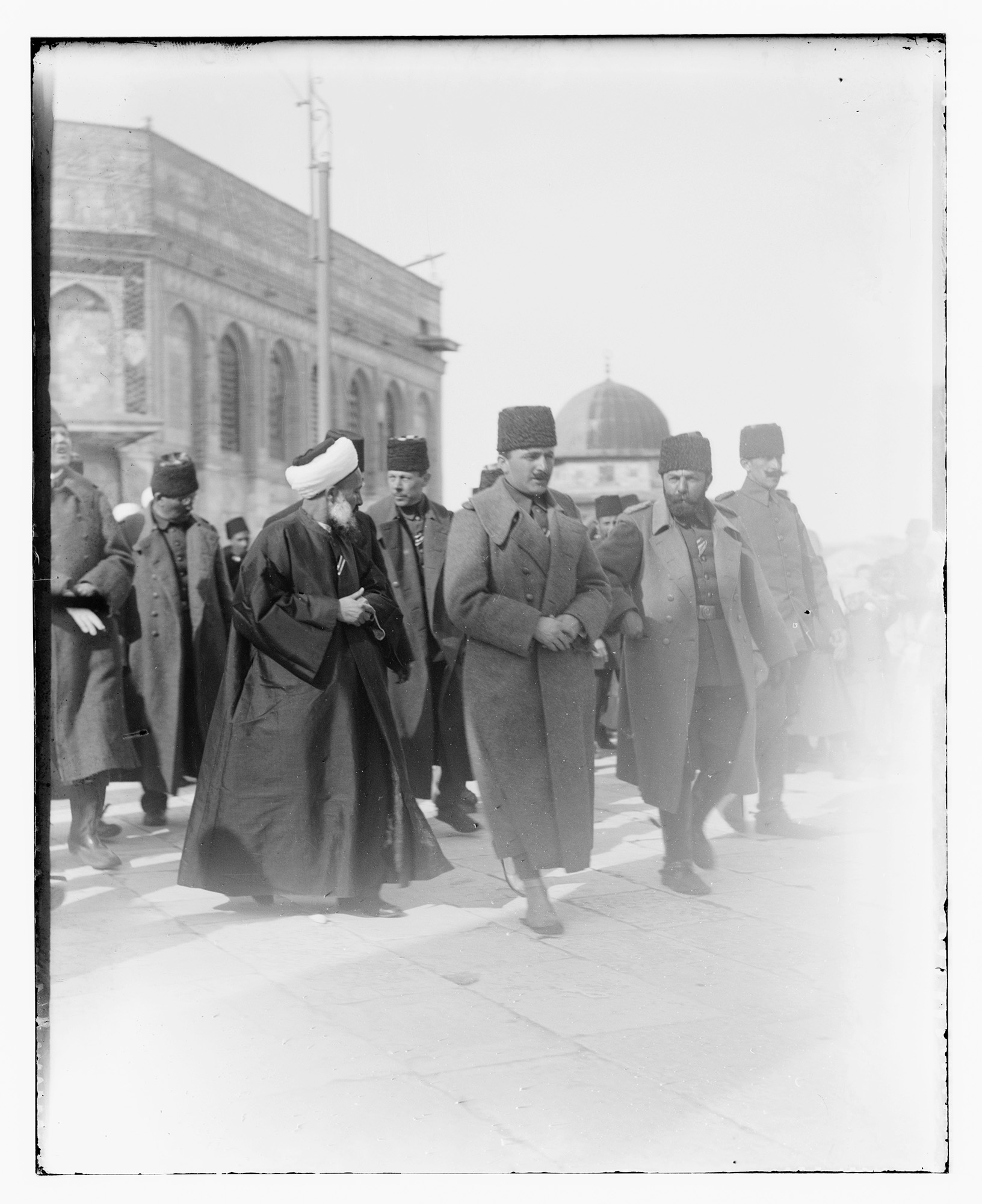 Black and white photograph of a group of men wearing heavy coats and fez hats walking away from a domed building.