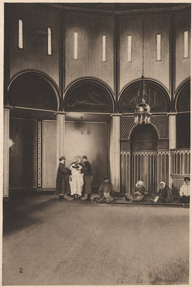 epia photograph of the interior of a mosque with repeated round arches. Three men are standing in discussion with each other while four other men are kneeling on mats on the ground.
