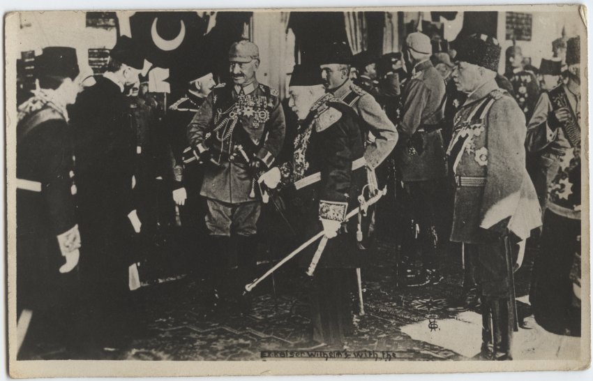 Black and white photograph of a man wearing a heavily decorated German uniform and steel helmet standing next to a man wearing a decorated robe and fez hat. Both are surrounded by various men in German and Ottoman uniforms.