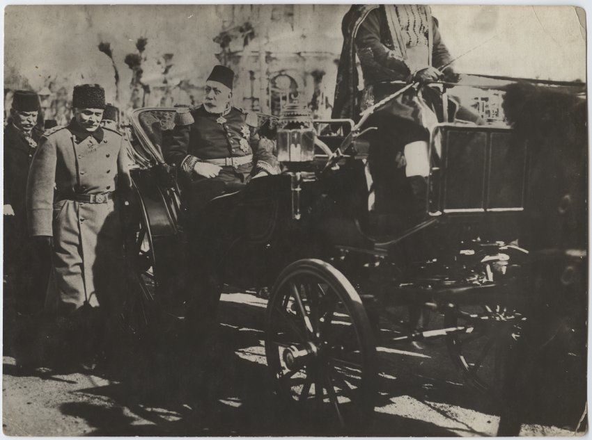 Black and white photograph of an older man wearing a fez hat sitting in an early automobile while other people walk behind the vehicle