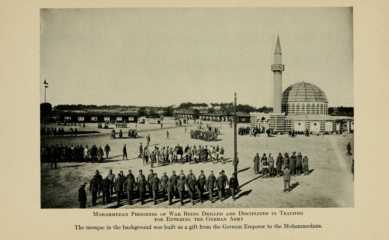 Black and white photograph of groups of men in prison uniform marching in lines across an open space. A mosque is in the background. Text: Mohammedan Prisoners of War Being Drilled and Disciplined in Training for Entering the German Army. The mosque in the background was build as a gift from the German Emperor to the Mohammedans.