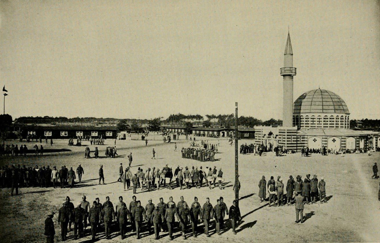 Black and white photograph of groups of men in prison uniform marching in lines across an open space. A mosque is in the background.