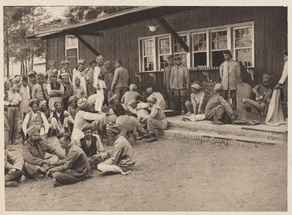 Sepia photograph of a large group of men sitting and standing in front of a low wooden building.