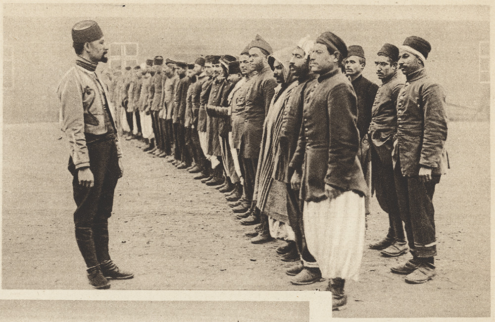 Sepia photograph of a man wearing a fez hat facing two lines of men standing at attention, wearing a variety of national clothing.