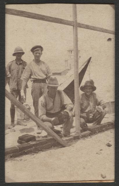 Sepia photograph of four men wearing wide-brimmed hats. Two are standing behind the other two, who are crouching and laying bricks along a wooden building frame.