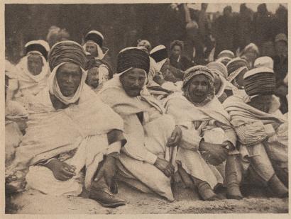 Sepia photograph of a group of darker-skinned men sitting on the ground wearing robes and head coverings.