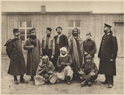 Sepia photograph of a group of men sitting and standing while posed for the photographer. They are wearing a mix of clothing from various nations.