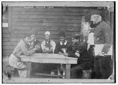 Black and white photograph of a small group of men smoking while playing cards around a wooden table outside a wooden building