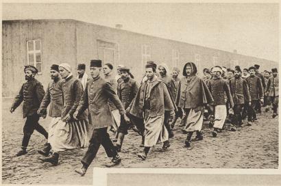 Sepia photograph of a column of men wearing coats walking out of the frame. Besides the coats, they are wearing a variety of trousers, robes, fez hats and turbans.