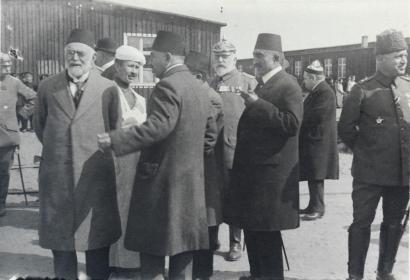 Black and white photograph of a group of men in suits and fez hats standing outside low wooden buildings