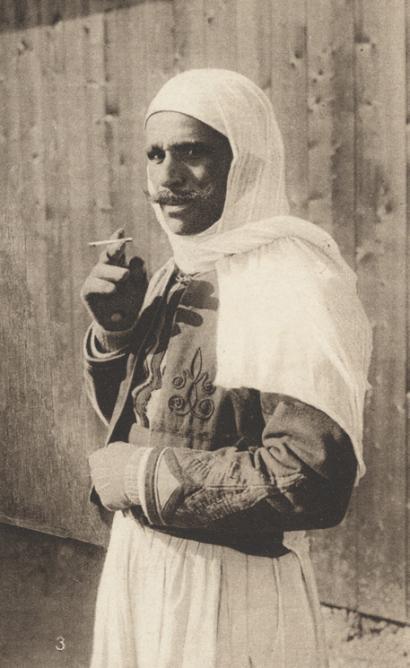 Sepia photograph of a mustached man wearing loose clothing and a flowing head covering, holding a cigarette in his fingers
