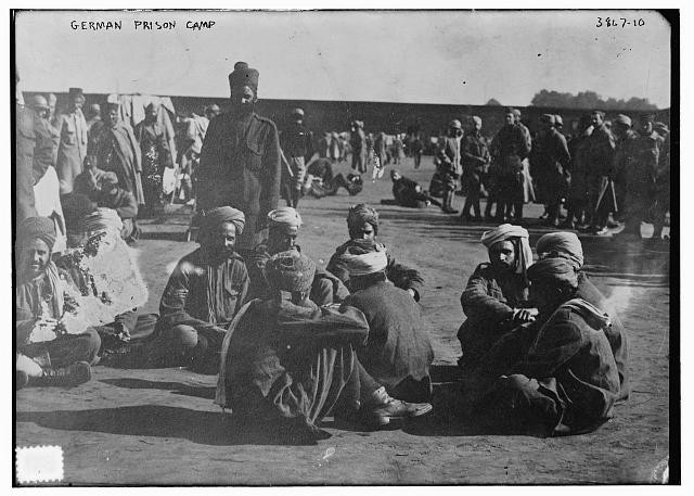 Black and white photograph of darker-skinned men dressed in loose clothing and turbans sitting and standing in groups in an open area.