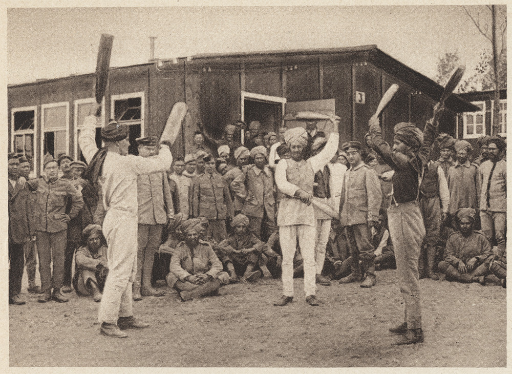 Sepia photograph of three men wearing turbans standing facing each other while each raising or waving two wooden paddles, in front of a large group of onlookers.
