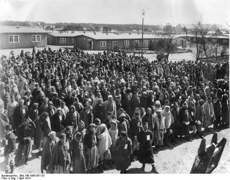 Black and white photograph of rows of men standing in an open space with low wooden buildings in the background