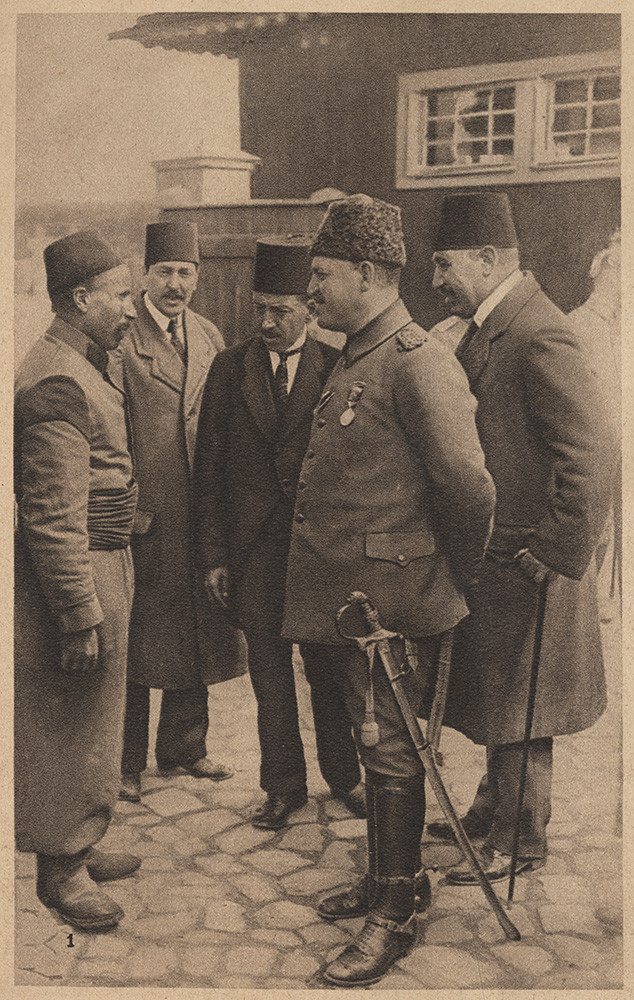 Sepia photograph of a man in military uniform with a fez hat and a sword speaking to a man wearing simple robes and a fez hat. Several other men in suits and fez hats stand around them.